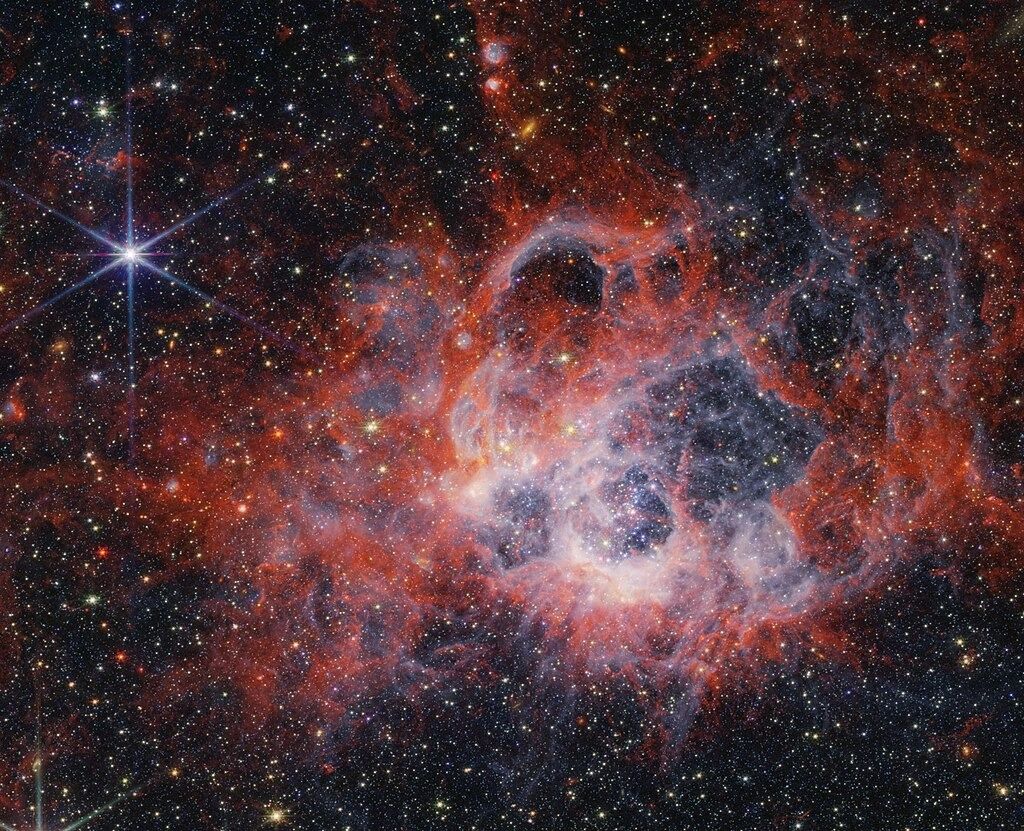 На этом снимке области звездообразования NGC 604, полученном с помощью NIRCam (камеры ближнего инфракрасного диапазона) космического телескопа NASA/ESA/CSA James Webb, показано, как звездные ветры от ярких, горячих молодых звезд прорезают полости в окружающем газе и пыли. Яркие оранжевые полосы на этом снимке указывают на присутствие молекул на основе углерода, известных как полициклические ароматические углеводороды, или ПАУ.